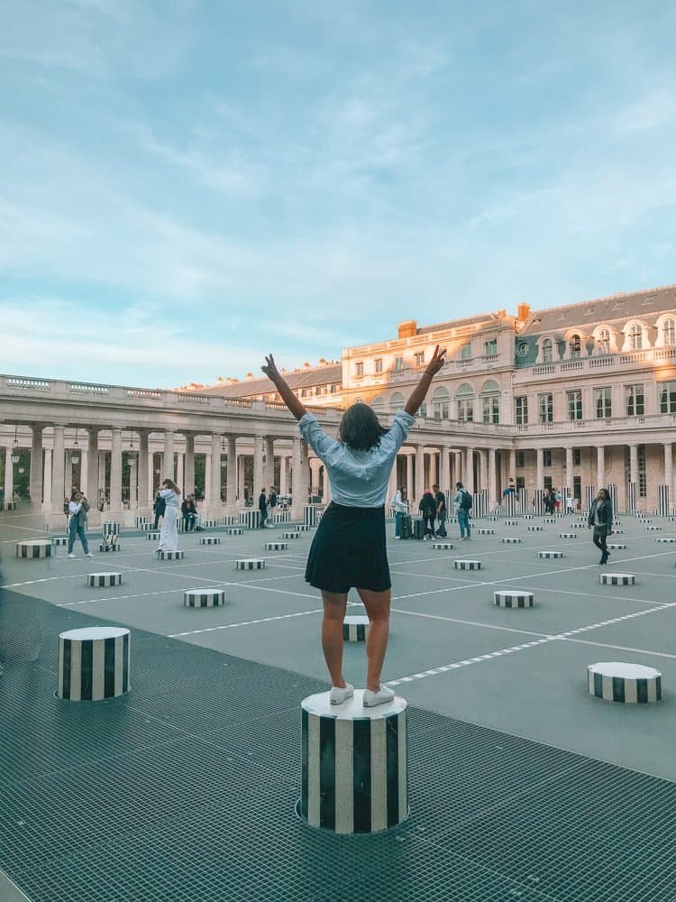 Kat's back to the camera as she has her hands up with peace fingers on a column at Palais Royale