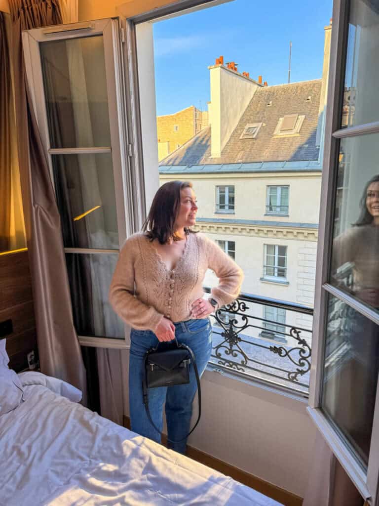 Kat posing at her hotel window with a sweater, jeans, and black purse on