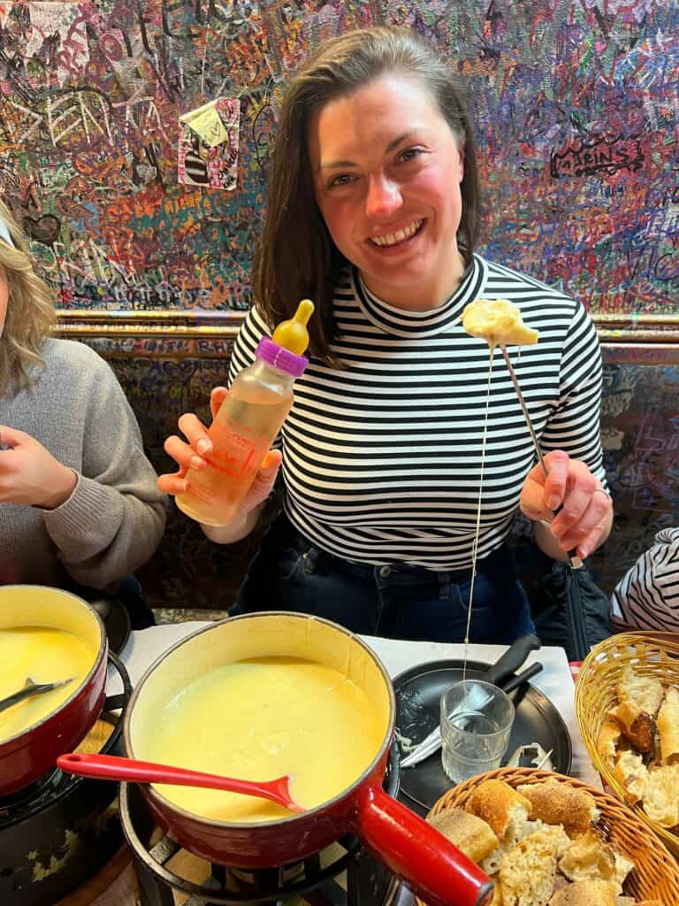 Kat with a baby bottle in one hand and fondue fork with baguette and fondue on it in the other at Le Refuge des Fondus
