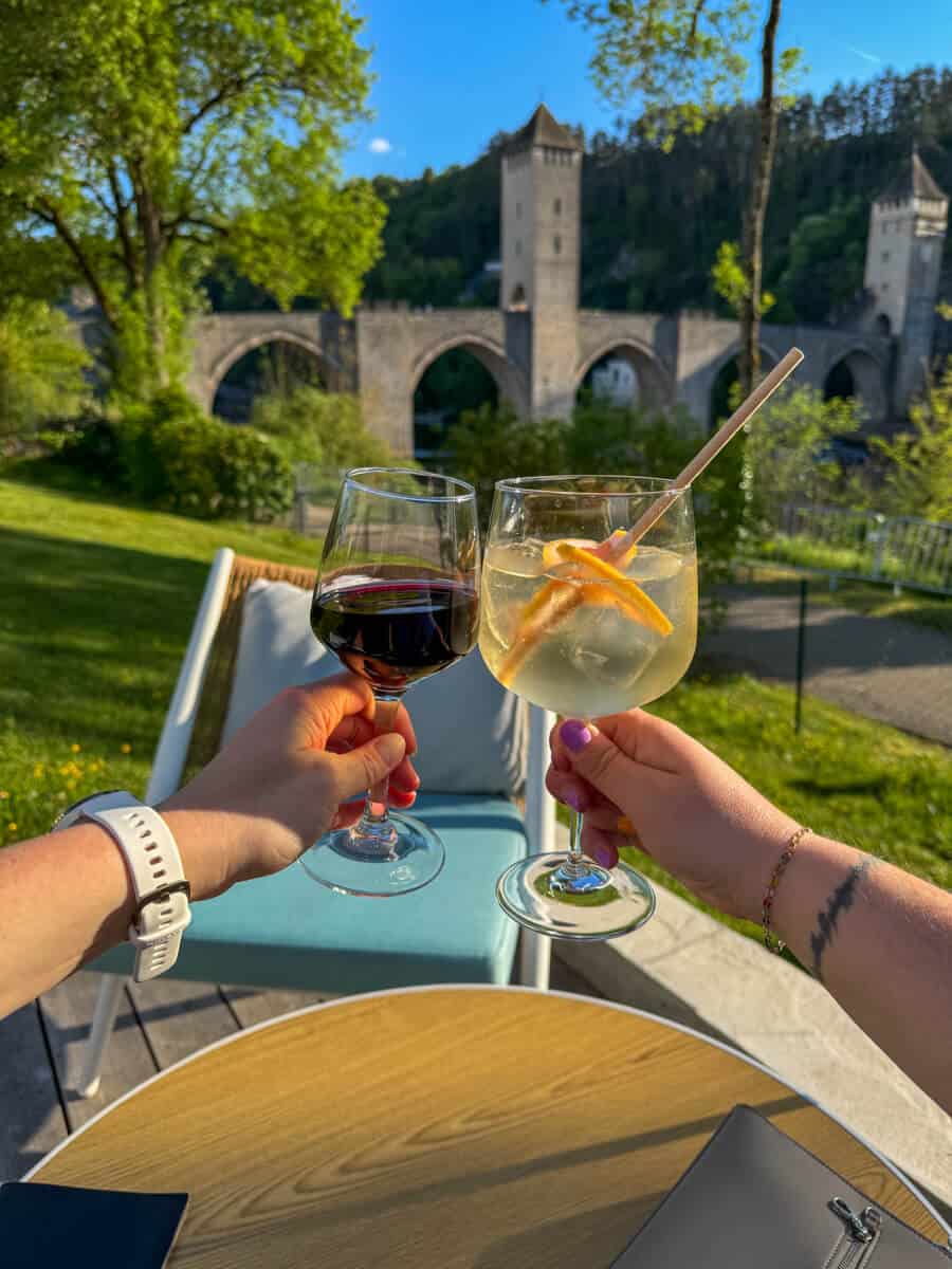 Wine and spritz drinks being held by hands in front of Cahors bridge